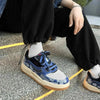 A person sits on stairs wearing black sweatpants, white socks, and Vexlly's Colorblock Patterned Sneakers in Blue. One hand rests on their knee, showcasing a silver bracelet. The setting appears to be outdoors.