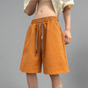 A person is standing against a plain backdrop, wearing a light beige shirt that's slightly open at the midriff, paired with Vexlly's oversized textured orange drawstring street-style shorts for men. These shorts, available in Asian sizes and designed for a relaxed fit with side pockets, are perfectly tailored according to the size chart.