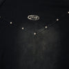 The black leather-like surface of the Punk Rivet T Shirts Patchwork Tee Top by Vexlly showcases a symmetrical pattern of small metallic studs arranged in an inverted V shape. A silver oval emblem with an intricate design is positioned at the center. Please note that slight variations in the pattern's alignment may occur due to hand measurement.