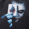 A stylized image features a mysterious figure holding a playing card, which displays a silhouette of Batman with the word "Joker" printed on it. The figure's face is shadowed, with piercing eyes and a faint smile, capturing the edgy allure typical of streetwear designs, reminiscent of Vexlly's Joker X DC Batman The Dark Knight '08 Heavy Graphic Tee.